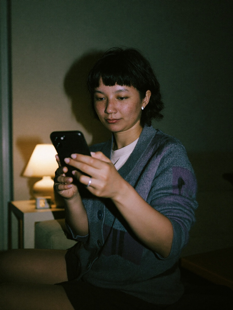 A mother in a dim bedroom at night, holding her sleeping toddler while checking her phone